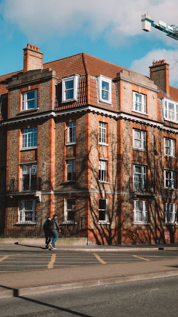 Photograph of a multi-storey residential building with several balconies, featuring pink exterior panels, glass and metal railings, and large windows. A patterned rug is hanging on one balcony railing